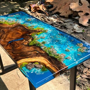 Puede incluir: Mesa rectangular de madera con un dise&ntilde;o &uacute;nico. La mesa presenta un patr&oacute;n de vetas de madera marr&oacute;n oscuro en un lado, que se transforma en una resina epoxi azul vibrante que imita una escena oce&aacute;nica. Los detalles verdes y beige se suman al tema costero.