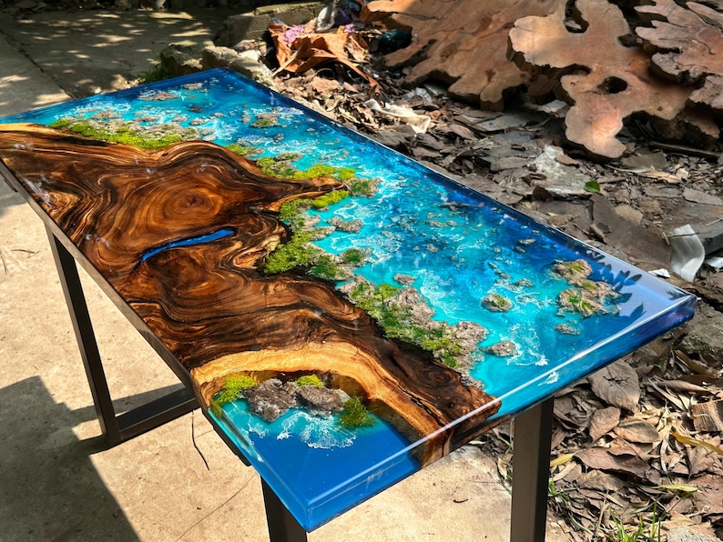 Puede incluir: Mesa rectangular de madera con un dise&ntilde;o &uacute;nico. La mesa presenta un patr&oacute;n de vetas de madera marr&oacute;n oscuro en un lado, que se transforma en una resina epoxi azul vibrante que imita una escena oce&aacute;nica. Los detalles verdes y beige se suman al tema costero.