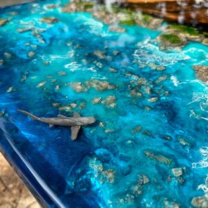 Puede incluir: Una obra de arte en resina que representa una escena oce&aacute;nica con tonos de azul y turquesa. La obra presenta un peque&ntilde;o tibur&oacute;n gris nadando en el agua, con peque&ntilde;as formaciones rocosas e islas verdes.