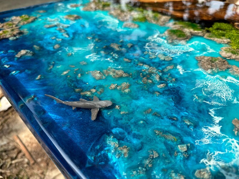 Puede incluir: Una obra de arte en resina que representa una escena oce&aacute;nica con tonos de azul y turquesa. La obra presenta un peque&ntilde;o tibur&oacute;n gris nadando en el agua, con peque&ntilde;as formaciones rocosas e islas verdes.