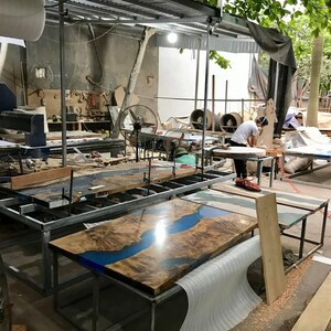 Puede incluir: Taller con varias mesas de madera en varias etapas de finalizaci&oacute;n. Las mesas presentan un dise&ntilde;o &uacute;nico con una incrustaci&oacute;n de resina epoxi azul en forma de r&iacute;o. Una persona est&aacute; trabajando en una mesa redonda.
