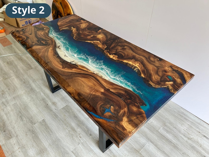 Puede incluir: Una mesa rectangular de madera con dise&ntilde;o de r&iacute;o. La mesa presenta una veta de madera marr&oacute;n oscuro con un r&iacute;o de resina azul y blanco que fluye por el centro. La mesa est&aacute; soportada por patas de metal negro. El texto "Style 2" est&aacute; en la esquina superior izquierda.