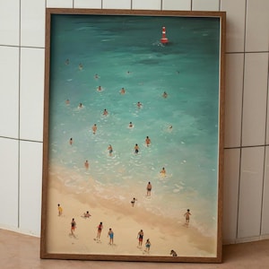 May include: A framed art print depicting a beach scene with people swimming in turquoise water. A red and white buoy floats in the distance. The sandy beach is populated with figures in various poses, suggesting a sunny day at the coast.
