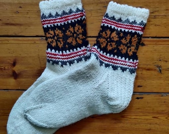 May include: A pair of hand-knitted socks with a cream-colored base and patterned cuffs. The cuffs feature a repeating design in black, orange, red, and gray. The socks are displayed on a wooden surface.