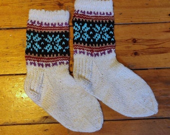 May include: A pair of hand-knitted socks featuring a cream-colored base with a decorative cuff pattern. The cuff displays horizontal bands of purple, brown, and black, accented with a bright blue geometric snowflake-like design on a black background.