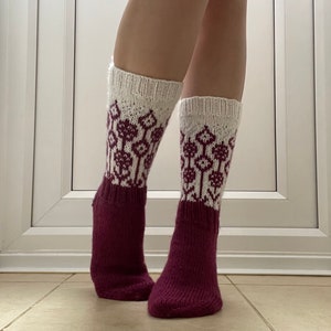 May include: A pair of knitted socks with a white cuff and a burgundy foot and leg. The white cuff features a repeating floral pattern in burgundy. The socks are knee-high and appear to be handmade.