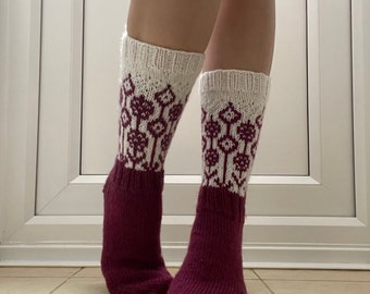 May include: A pair of knitted socks with a white cuff and a burgundy foot and leg. The white cuff features a repeating floral pattern in burgundy. The socks are knee-high and appear to be handmade.