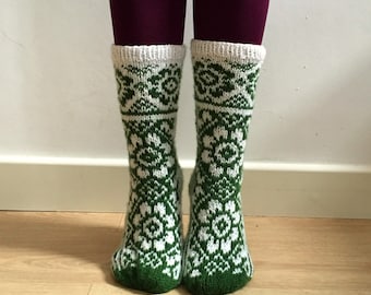 May include: A pair of green and white patterned socks. The socks feature a repeating floral design and a white cuff. The socks are worn with dark purple leggings and are displayed on a light wood floor.
