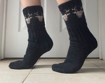 May include: A pair of black knit socks with a patterned design in tan and white at the top. The socks are thick and appear to be handmade. The socks are worn on a person's feet.