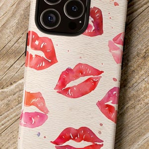 Könnte beinhalten: Ein Handyhülle mit einem Muster aus roten und rosa Lippen auf cremefarbenem Hintergrund. Die Hülle liegt auf einer Holzoberfläche, mit einer Kamera und einer Pflanze im Hintergrund.