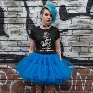 Könnte beinhalten: Ein schwarzes T-Shirt mit einer Grafik einer Kreatur und Hunden, kombiniert mit einem leuchtend blauen Tütü. Der Text "WorstCaseScenario" befindet sich unter der Grafik. Das Outfit wird durch schwarze Stiefel und Netzstrümpfe ergänzt, vor einer Backsteinmauer mit Graffiti.