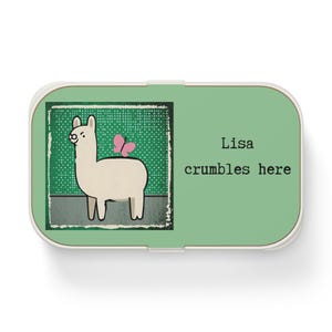 Könnte beinhalten: Eine rechteckige Dose mit weißem Rand und grünem Deckel. Der Deckel zeigt ein Cartoon-Lama mit einem rosa Schmetterling und dem Text "Lisa crumbles here" in Schwarz.