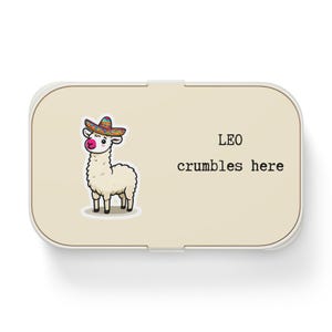 Könnte beinhalten: Eine beige Lunchbox mit weißem Rand. Die Lunchbox zeigt ein Cartoon-Lama mit einem bunten Sombrero. Der Text auf der Lunchbox lautet "LEO crumbles here."