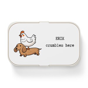 Könnte beinhalten: Weiße Brotdose mit einer Cartoon-Illustration eines braunen Dackelhundes mit einem weißen Huhn auf dem Rücken. Der Text "ERIK crumbles here" ist in Schwarz gedruckt.