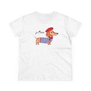 Könnte beinhalten: Weißes T-Shirt mit einem Cartoon-Dackel, der ein rotes Barett und einen Schal sowie ein blau-weiß gestreiftes Hemd trägt. Der Satz "je t'aime" steht in schwarzer Schrift über dem Hund.