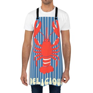 Könnte beinhalten: Schürze mit blau-weißem Streifenmuster und einer großen roten Hummerillustration. Das Wort "DELICIOUS" ist unten in Weiß aufgedruckt. Die Schürze hat schwarze Träger und Knöpfe.