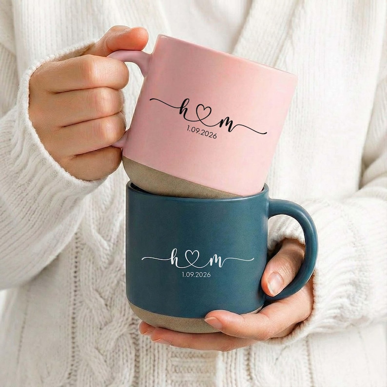 May include: Two ceramic mugs, one pink and one teal, stacked together. Each mug features a white script design with the letters "h♡m" and the date "1.09.2026". The mugs have a light brown base and are held by a person wearing a white sweater.
