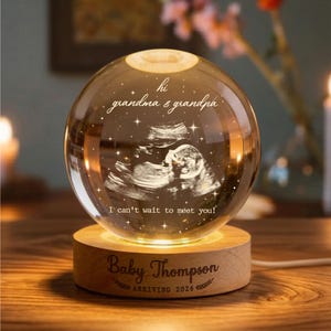 May include: A glowing crystal ball with a sonogram image inside, resting on a wooden base. The text "hi grandma & grandpa" and "I can't wait to meet you!" are inscribed on the ball. The base reads "Baby Thompson ARRIVING 2026".