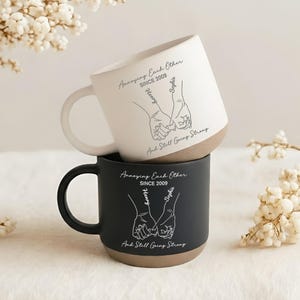 May include: A white ceramic mug stacked on a black ceramic mug, both featuring a line art illustration of two hands holding pinky fingers. The text reads Annoying Each Other SINCE 2009, Henry, Sophie, and And Still Going Strong in a script font.