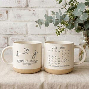 May include: Two off-white ceramic mugs with a speckled finish and a tan base. One mug features the letters S and J connected by a heart, the names Sarah & Jacob, and 35th Wedding Anniversary 1991-2026. The other shows an October 1991 calendar.