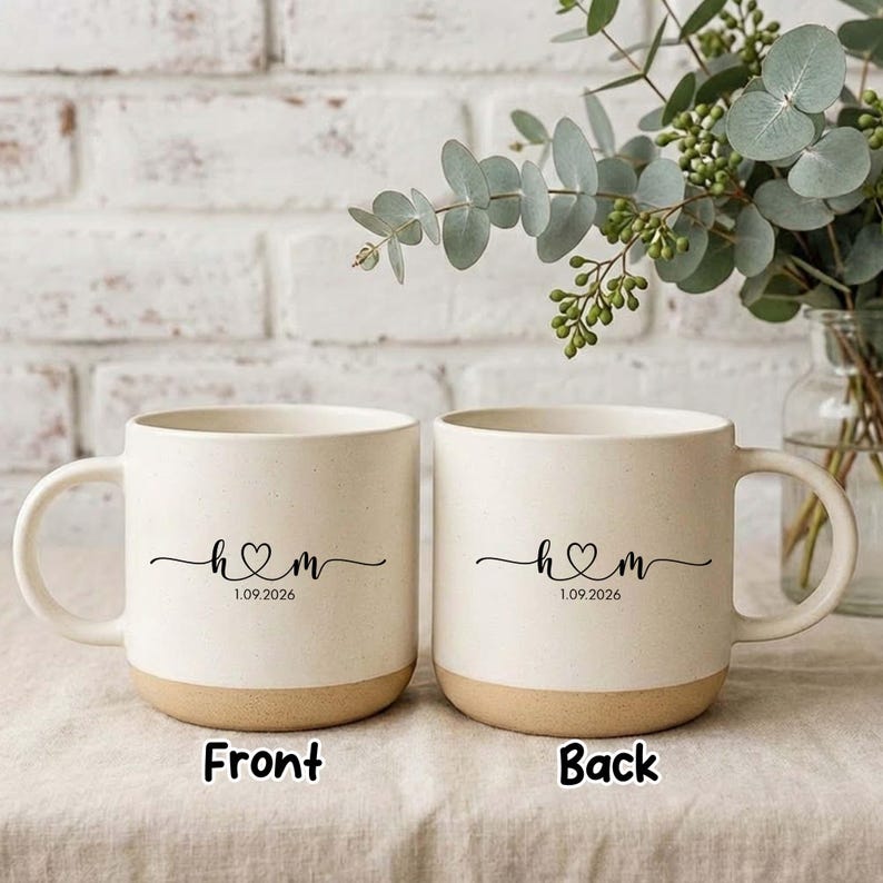 May include: Two off-white ceramic mugs with a tan base. Each mug features a black heart design with the letters "H & M" and the date "1.09.2026". The mugs are on a light-coloured surface with greenery in the background.
