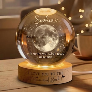 May include: A clear glass sphere lamp featuring a laser-etched moon and stars inside. The text "Sophia" and "THE NIGHT YOU WERE BORN 09.08.1998" are etched on the sphere. The light sits on a wooden base engraved with "I LOVE YOU TO THE MOON AND BACK".