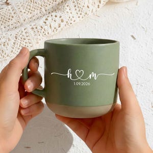 May include: A green ceramic mug with a tan base is held by two hands. The mug features white text: "h♡m" above the date "1.09.2026". The mug has a curved handle and a matte finish.