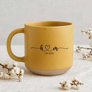 May include: Mustard yellow ceramic mug with a speckled finish and a light brown base. The mug features a black script design with the initials "h & m" connected by a heart, and the date "1.09.2026".