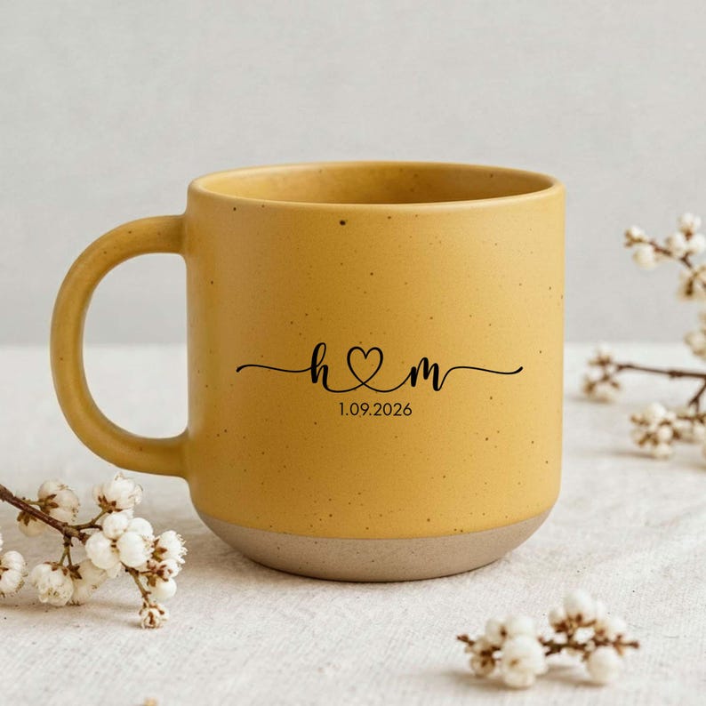 May include: Mustard yellow ceramic mug with a speckled finish and a light brown base. The mug features a black script design with the initials "h & m" connected by a heart, and the date "1.09.2026".