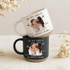 May include: Two ceramic mugs, one white and one black, stacked. Each mug features a photo of a couple embracing, with the text "You Will Always Be" and "My Greatest Adventure." The white mug has a beige base and handle, while the black mug has a black handle and beige base.