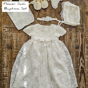 Puede incluir: Un conjunto de bautizo de encaje blanco, que incluye un vestido, gorro, zapatos, calcetines, diadema y faja. El vestido presenta un dise&ntilde;o floral y una cinta de sat&eacute;n. El conjunto se muestra sobre una superficie de madera. El texto en la imagen dice "Flower Sash Baptism Set."