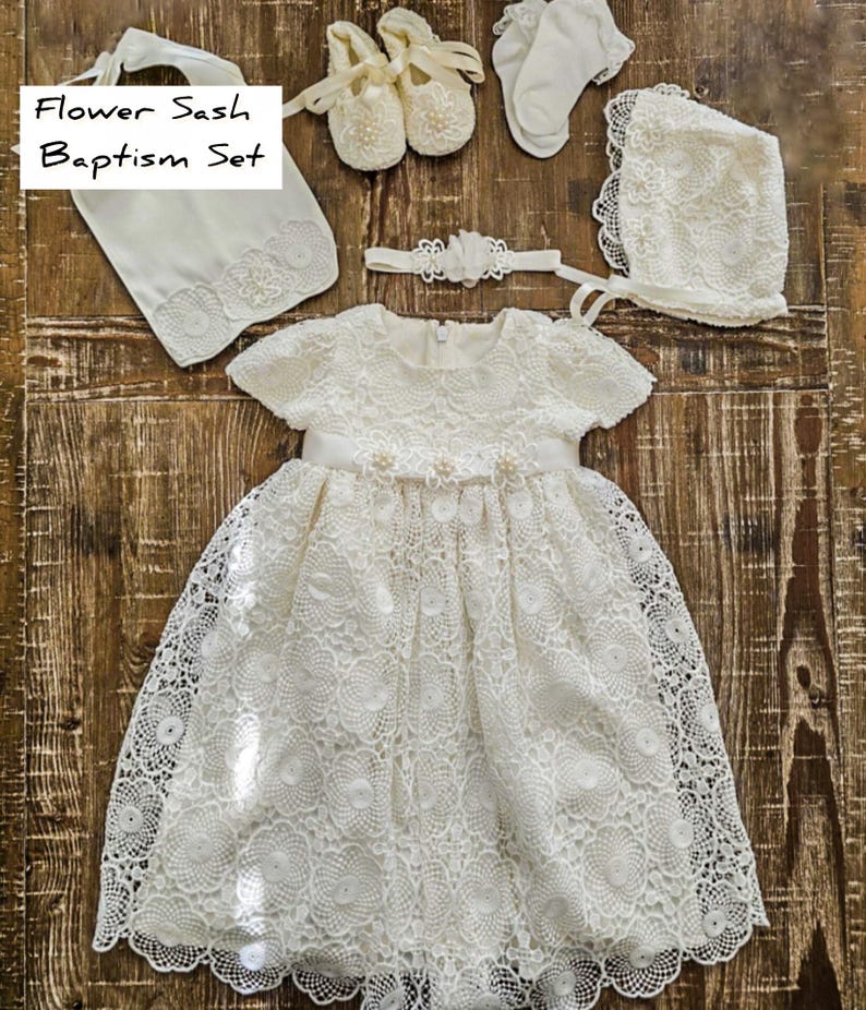 Puede incluir: Un conjunto de bautizo de encaje blanco, que incluye un vestido, gorro, zapatos, calcetines, diadema y faja. El vestido presenta un dise&ntilde;o floral y una cinta de sat&eacute;n. El conjunto se muestra sobre una superficie de madera. El texto en la imagen dice "Flower Sash Baptism Set."