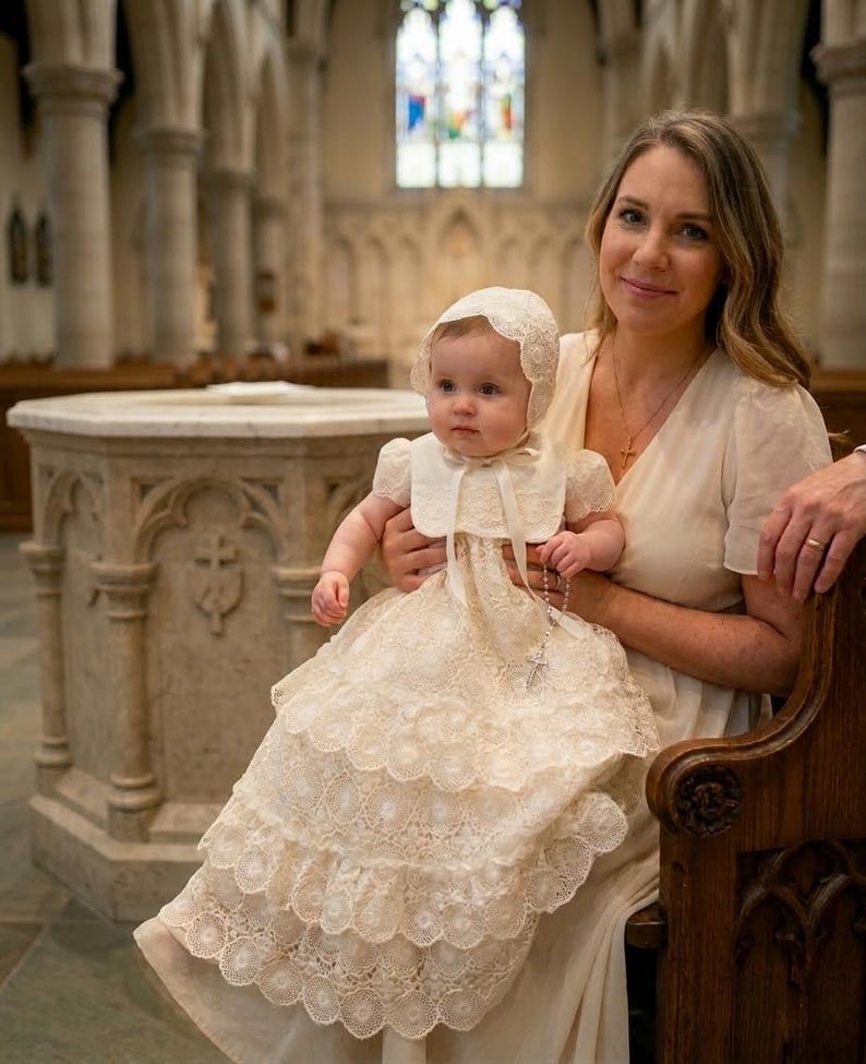 Puede incluir: Una beb&eacute; con un vestido de bautizo y un gorro de encaje color crema es sostenida por una mujer con un vestido crema. El vestido tiene m&uacute;ltiples capas de encaje y un cuello a juego. El fondo muestra el interior de una iglesia con vidrieras.