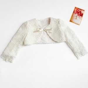 Puede incluir: Bolero de encaje color marfil con mangas largas y un peque&ntilde;o lazo en la parte delantera. La chaqueta est&aacute; hecha de un delicado encaje con estampado floral. Una peque&ntilde;a tarjeta con un dise&ntilde;o floral est&aacute; en el fondo.