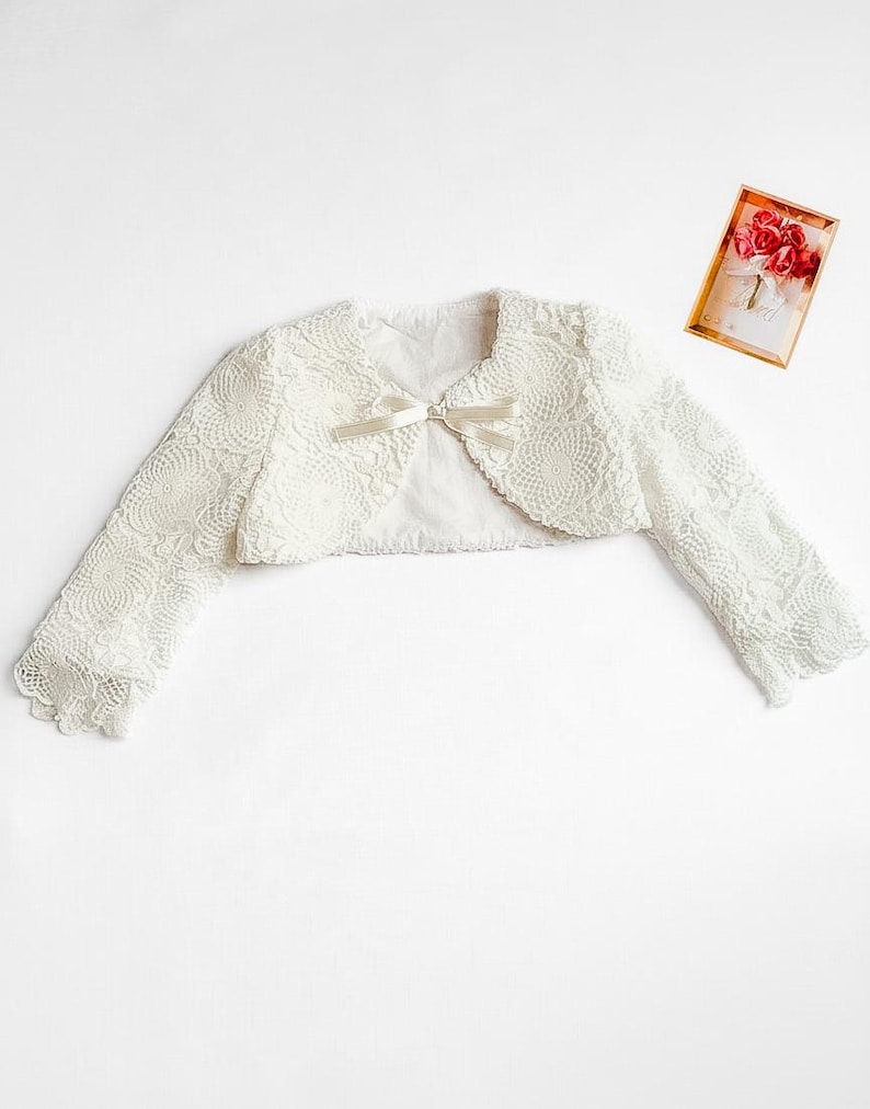Puede incluir: Bolero de encaje color marfil con mangas largas y un peque&ntilde;o lazo en la parte delantera. La chaqueta est&aacute; hecha de un delicado encaje con estampado floral. Una peque&ntilde;a tarjeta con un dise&ntilde;o floral est&aacute; en el fondo.