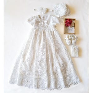 Puede incluir: Un vestido de bautizo de encaje blanco con gorro, diadema y patucos a juego. Tambi&eacute;n se incluye un peque&ntilde;o marco de madera con rosas rojas y la palabra "Loved". El conjunto es para un beb&eacute;.