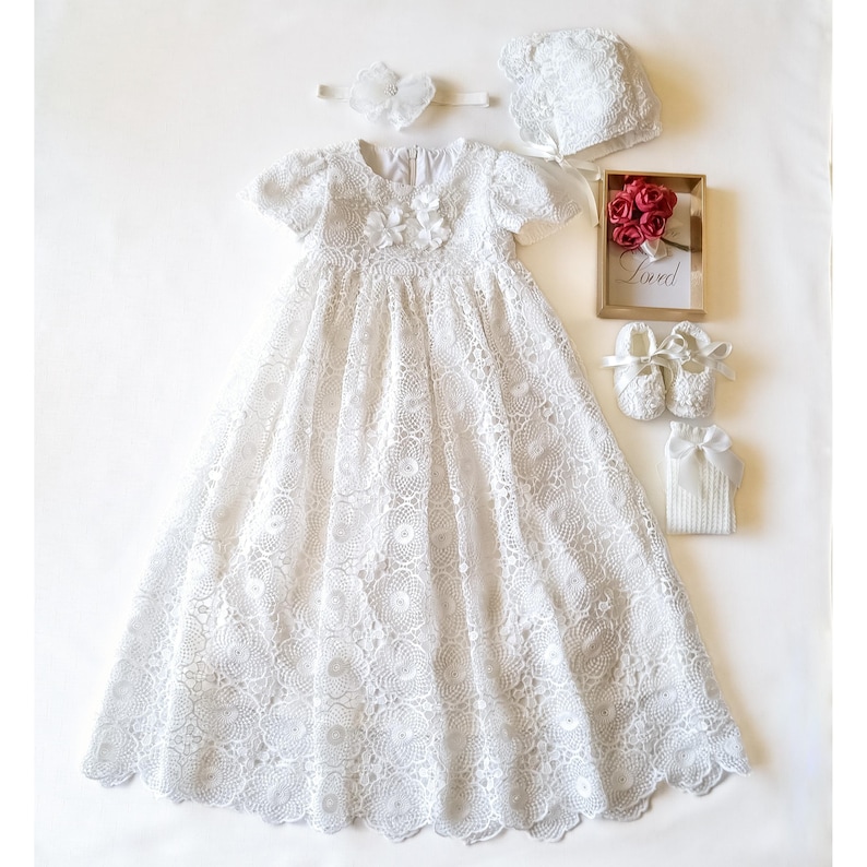 Puede incluir: Un vestido de bautizo de encaje blanco con gorro, diadema y patucos a juego. Tambi&eacute;n se incluye un peque&ntilde;o marco de madera con rosas rojas y la palabra "Loved". El conjunto es para un beb&eacute;.