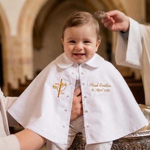 Op de afbeelding: Een lachende baby draagt een witte doopcape met een gouden geborduurd kruis- en duivenontwerp. De cape heeft een Peter Pan-kraag en is aan de voorkant dichtgeknoopt. De naam en datum van de baby zijn op de cape geborduurd.