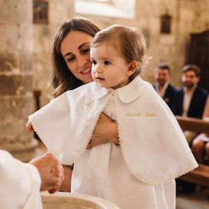 Puede incluir: Un vestido de bautizo blanco que incluye una capa corta con cuello redondo y ribete de encaje festoneado. La capa está bordada con el nombre "Marielle Stella" en hilo dorado a la izquierda y un pequeño emblema dorado a la derecha.