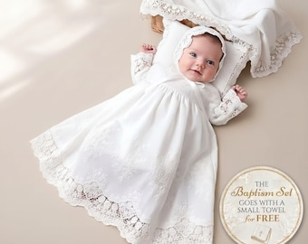 May include: A white infant baptism gown featuring long sleeves with lace cuffs, a matching bonnet, and a full skirt with intricate floral embroidery and a wide lace hem. The set includes a white pillow and a decorative cloth with lace trim.