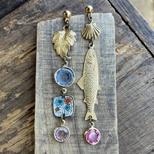 May include: A pair of mismatched gold-toned earrings. One earring features a leaf, a blue gem, a floral square, and a pink gem. The other earring has a seashell and a fish pendant with a pink gem. The earrings are on a wooden surface.