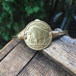 Op de afbeelding: Een goudkleurige armband met een vintage Buffalo-nikkel uit de Verenigde Staten. De munt, met de woorden "United States of America" en "Five Cents", is het middelpunt. De band van de armband is gemaakt van goudkleurig draad, gewikkeld rond de munt.