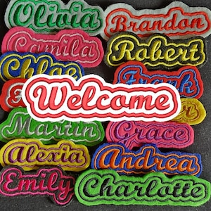 Può includere: Una collezione di toppe di stoffa ricamate colorate con nomi come Olivia, Brandon, Robert e altri. Le toppe sono disposte in una pila, con una grande toppa rossa che dice "Benvenuti" al centro.