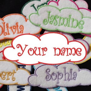 Puede incluir: Una colección de parches en forma de nube de fieltro blanco con nombres bordados, incluyendo Olivia, Jasmine, Robert, Sophia y un parche en blanco que dice "Your name".