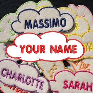 Puede incluir: Una colección de parches de fieltro en forma de nube de colores con nombres bordados, incluyendo "Massimo", "Your Name", "Charlotte", "Sarah" y "Ryan".