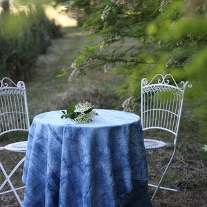 May include: A blue tie-dye tablecloth with white accents covers a round table with two white metal chairs. A small bouquet of white flowers sits on the table.