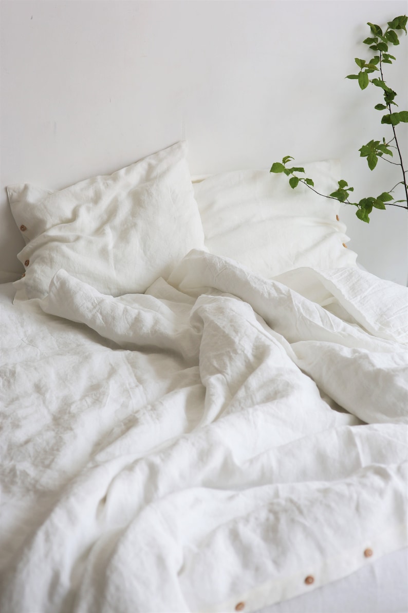 Puede incluir: Ropa de cama de lino blanco con un edred&oacute;n arrugado y dos fundas de almohada blancas. La ropa de cama est&aacute; sobre un marco de cama blanco y hay una planta verde en el fondo.