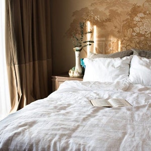 Puede incluir: Una cama de lino blanco con un libro en la colcha. La cama est&aacute; hecha con almohadas blancas y una funda n&oacute;rdica blanca. La cabecera est&aacute; contra una pared con un patr&oacute;n de papel tapiz floral.