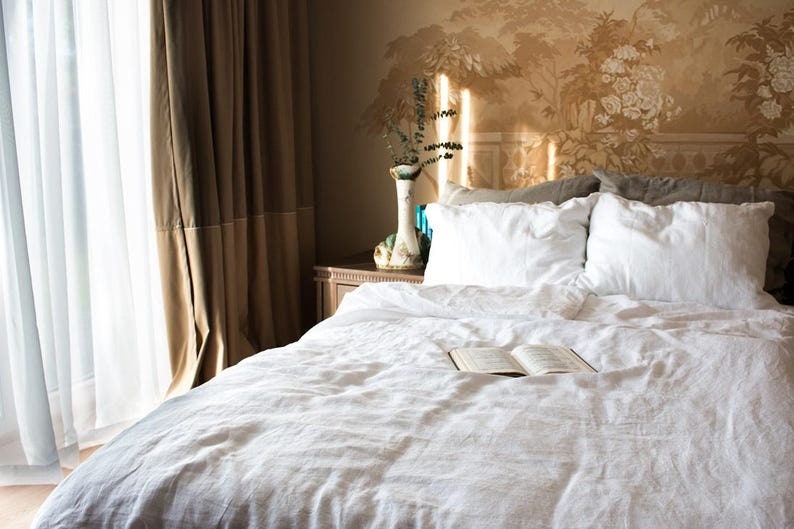 Puede incluir: Una cama de lino blanco con un libro en la colcha. La cama est&aacute; hecha con almohadas blancas y una funda n&oacute;rdica blanca. La cabecera est&aacute; contra una pared con un patr&oacute;n de papel tapiz floral.