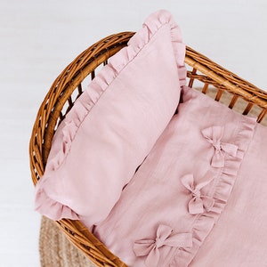 May include: A pink linen baby bedding set with a pillow and a fitted sheet. The bedding has a ruffled edge and bows on the sheet.